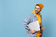 © ViDi Studio - Side view of young hipster guy in fashion jeans denim clothes posing isolated on pastel blue background in studio. People lifestyle concept. Mock up copy space. Hold laptop pc computer, looking back.