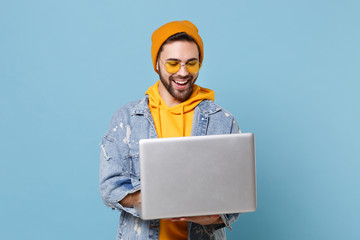 Funny young hipster guy in fashion jeans denim clothes posing isolated on pastel blue background studio portrait. People emotions lifestyle concept. Mock up copy space. Working on laptop pc computer.