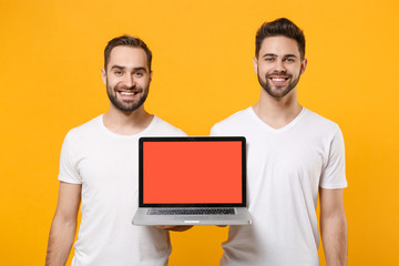 Wall Mural - Smiling young men guys friends in white t-shirts posing isolated on yellow orange background in studio. People lifestyle concept. Mock up copy space. Hold laptop pc computer with blank empty screen.
