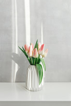 Light Pink Tulips In A White Geometric Ceramic Vase Stand On A White Table Near Grey Wall. Bouquet Of Flowers In The Morning Sun Beams.