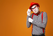Pensive Mime In Red Hat, White Gloves And Striped T-shirt On Blank Orange Background