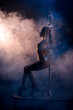 side view of attractive stripper in fishnet tights dancing striptease near pylon on blue with smoke