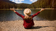 Back view of female tourist in hat with hands up enjoying freedom and amazing scenery of mountain lake.