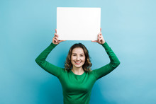 Curly Girl Wearing In Green Sweater Holding A White Board Copy Space Above Her Head Against Blue Background