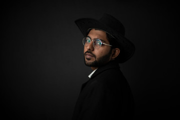 Fashion portrait of young and handsome Indian Bengali brunette man with striped formal shirt, black suit, glasses and cowboy hat in black copy space background. Indian lifestyle and fashion photograph