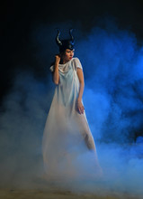 Beautiful Girl In A Black Helmet With Horns And In White Clothes On A Blue Background In Smoke