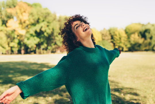 Horizontal image of happy young woman smiling posing against nature background with windy curly hair, have positive expression, wearing in green sweater. People, travel and lifestyle concept.