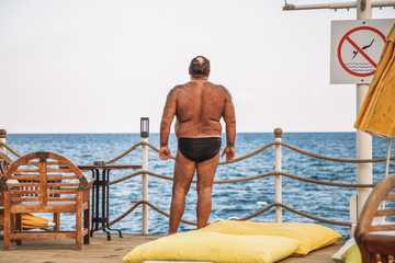 Adult hairy man standing at sea beach sunbathing looking at beautiful blue sea water. Horizontal color photography.