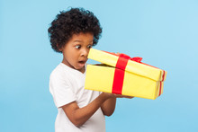 Portrait Of Amazed Curious Adorable Little Boy Peeking Inside Gift Box, Unpacking Present With Funny Astonished Expression, Impatient Child Unboxing Birthday Surprise. Studio Shot Blue Background