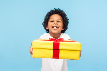 Take it! Full of joy amazing cute little boy with curly hair giving gift box to camera with excited smile, child greeting on holiday and sharing present. indoor studio shot isolated on blue background