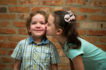 A little girl kisses a boy on the cheek. Brother and sister. Against a brick wall.