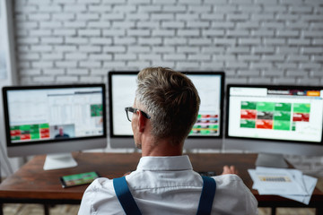 Where future and security collapse. Rear view of middle-aged trader sitting by desk in front of pc and analyzing stock market chart, while working in the office.