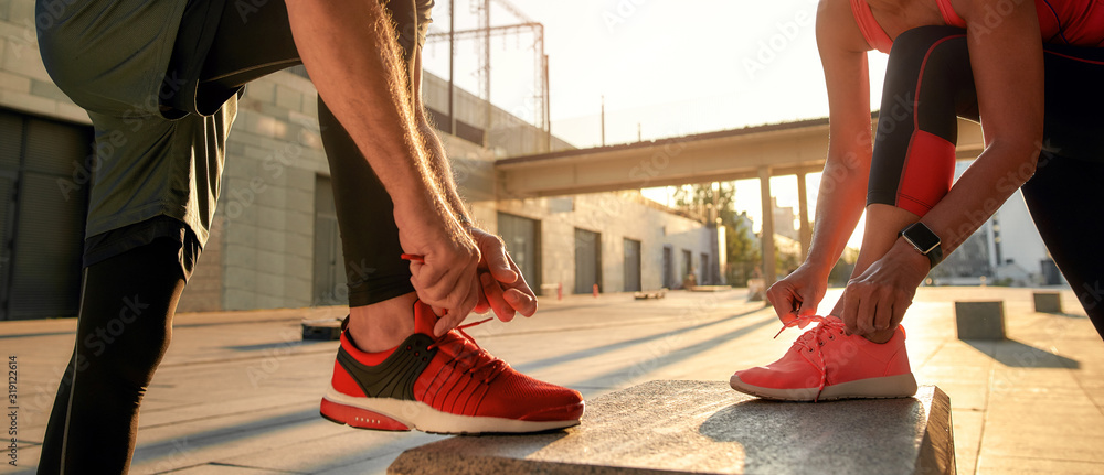 Active morning. Close up photo of two people in sport clothes tying shoelaces before running together outdoors - obrazy, fototapety, plakaty 