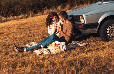 Wall Mural - Beautiful young couple enjoying picnic time on the sunset. They drinking tea and sitting in a meadow leaning on old fashioned car.