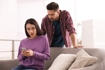 Wall Mural - Distrustful young man peering into girlfriend's smartphone at home