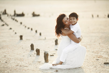 Mother and son is sitting on the sand and hugging, dressed in white clothes