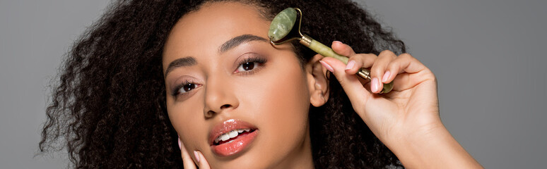 Wall Mural - panoramic shot of tender african american girl using jade roller, isolated on grey