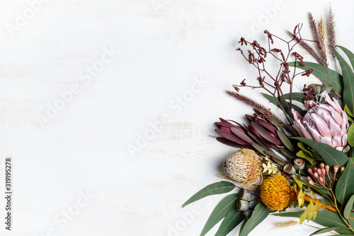Beautiful flat lay floral arrangement of mostly Australian native flowers, including protea, banksia, kangaroo paw eucalyptus leaves and gum nuts on a white background.