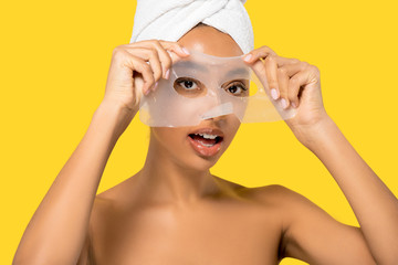 Wall Mural - portrait of shocked african american girl with towel on head applying hydrogel mask, isolated on yel