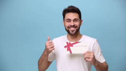 Wall Mural - Smiling unshaven bearded young guy 20s wearing white t-shirt isolated on pastel blue background in studio. People sincere emotions lifestyle concept. Pointing on gift certificate card coupon voucher