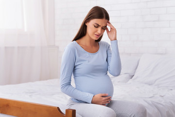 Wall Mural - Unhappy Expectant Girl Touching Head And Belly Sitting On Bed