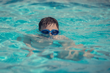 European boy in swimming goggles is swimming in the hotel’s pool during his summer vacations.