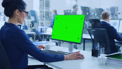 Over the Shoulder: Female Chief Engineer Working on Personal Computer, Display is Green Mock-up Screen or Chroma Key. Industrial Factory with High-Tech Machinery and Professional Workers