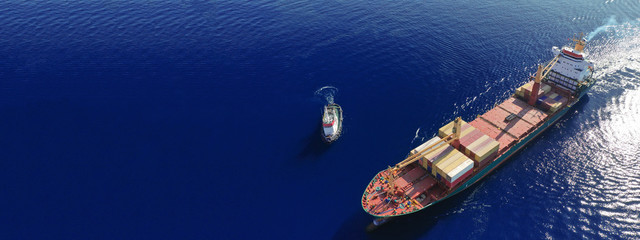 Aerial drone ultra wide top down photo of container ship cruising in Mediterranean port with deep blue sea assisted by tug boat