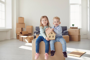 Two children are sitting on a box for moving they are holding a model of a house in a new bright room.