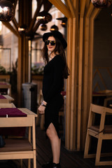 Photo of brunette lady posing on wooden background in cafe.Fashion style portrait. Girl wearing dark casual dress, sunglasses and dark hat .Fashion concept.