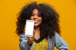 © Brastock Images - Portrait of cheerful, positive, attractive young woman in jeans shirt, having smart phone with white screen in hand, pointing with forefinger to product, isolated on yellow background