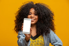 Portrait Of Cheerful, Positive, Attractive Young Woman In Jeans Shirt, Having Smart Phone With White Screen In Hand, Pointing With Forefinger To Product, Isolated On Yellow Background