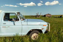 Old Blue Pickup Truck Close Up In An Open Field With Old Rundown Barn Shed And Blue Sky Cloud Background