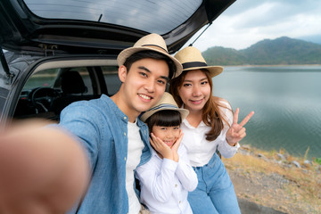 Portrait of Asian family sitting in car with father, mother and daughter selfie with lake and mountain view by smrtphone while vacation together in holiday. Happy family time.