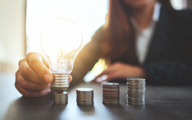 Wall Mural - businesswoman holding and putting lightbulb on coins stack on table for saving energy and money conc
