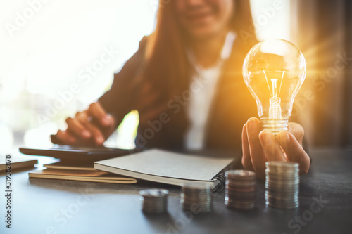 Fotomural Businesswoman holding a lightbulb with coins stack on table, saving energy and m