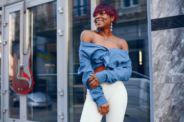 Wall Mural - beautiful and stylish dark-skinned girl with red short hair, dressed in a blue top with long sleeves and white pants standing near gray building