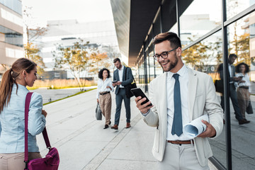 Wall Mural - Portrait of smiling businessman standing in front of modern office buildings looking at phone.