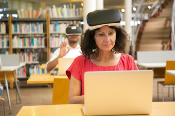 Front view of cheerful Caucasian woman setting VR headset. Smiling mature user with virtual reality glasses sitting at library. Technology concept