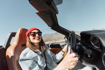 Happy woman in red hat driving convertible car while traveling on the desert road. Carefree lifestyle and travel concept