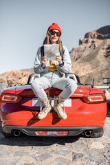 Wall Mural - Lifestyle portrait of a carefree woman dressed casually in jeans and red hat sitting with map on the car trunk, enjoying road trip on the desert valley