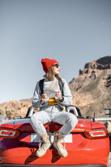 Wall Mural - Lifestyle portrait of a carefree woman dressed casually in jeans and red hat sitting with map on the car trunk, enjoying road trip on the desert valley