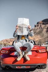 Lifestyle portrait of a carefree woman dressed casually in jeans and red hat sitting with map on the car trunk, enjoying road trip on the desert valley