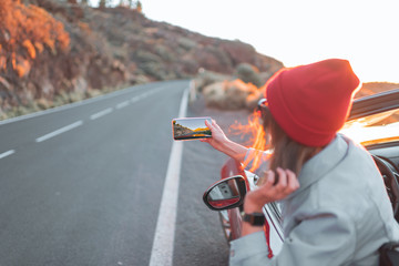 Young woman enjoying beautiful sunset view, leaning out of the car window with mobile phone. Carefree lifestyle and travel concept