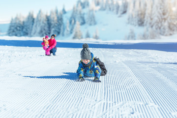 Boy falls into the snow while being watched by his mom and sister from the background. Concept of improving children's health through playing in the snow and clean mountain air