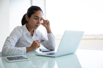 Concentrated young businesswoman using laptop in office. Focused young businesswoman sitting at desk and using laptop computer in modern office. Business and technology concept