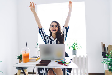 Cheerful young woman sitting at desk with raised hands