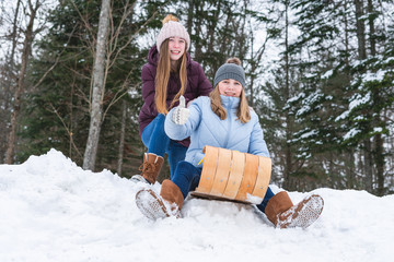  Two happy teen girls/friends sitting on a wood toboggan/sled while at the edge of a snow covered hill in a park and one girl holding her thumb up.