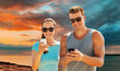 © Syda Productions - fitness, sport and healthy lifestyle concept - happy couple in sports clothes and sunglasses with smartphones over sea and sunset sky background