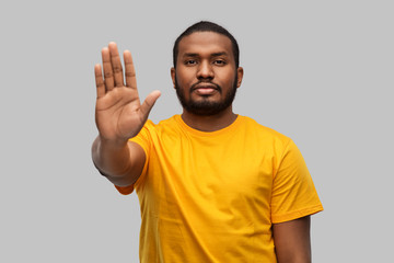 Wall Mural - warning and people concept - young african american man in yellow t-shirt showing stop gesture over grey background